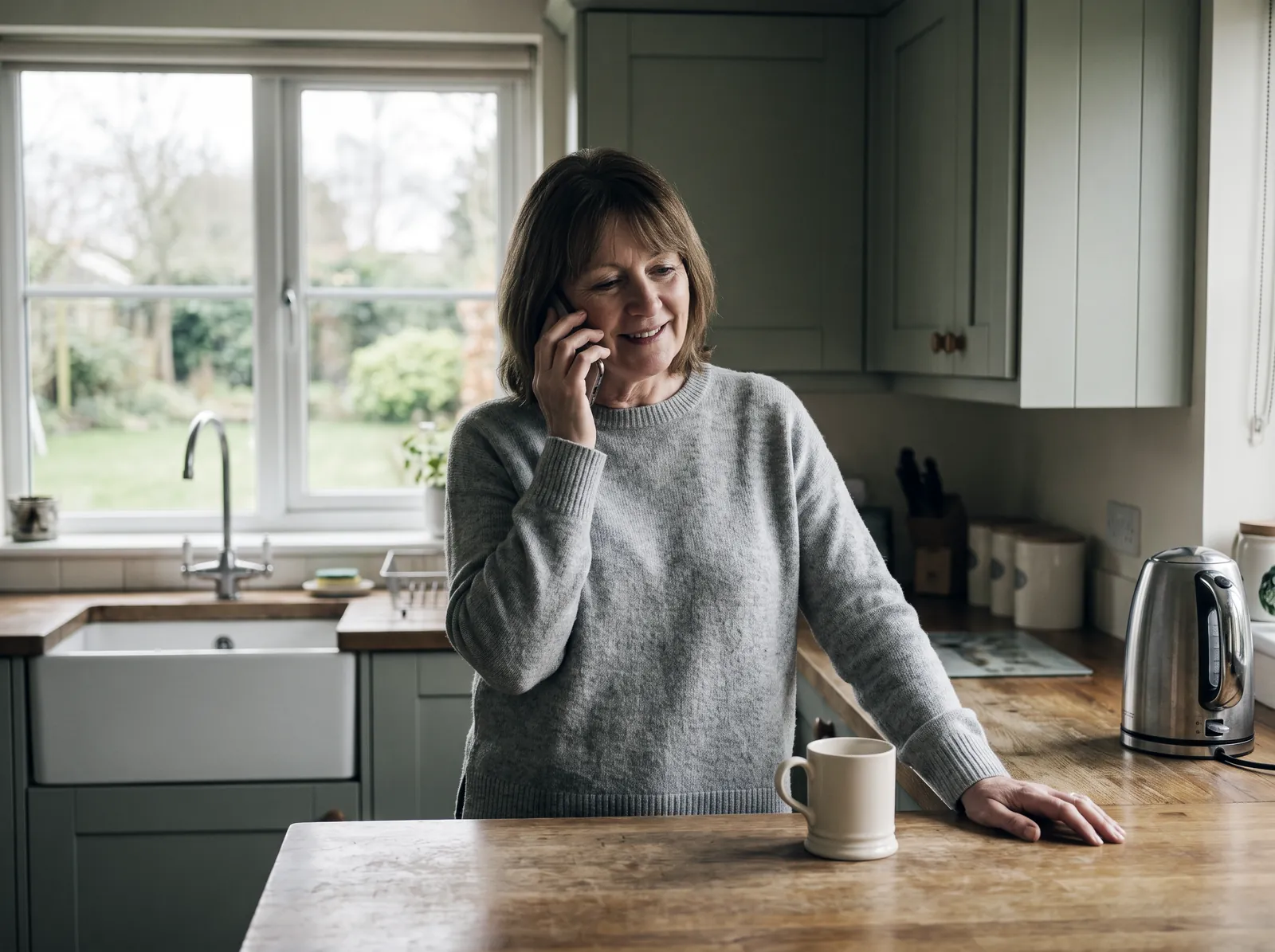 A woman on the phone in her kitchen with a mug of tea